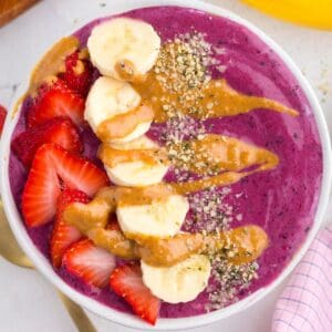 A smoothie bowl topped with sliced strawberries, banana, peanut butter drizzle, and sprinkled seeds.