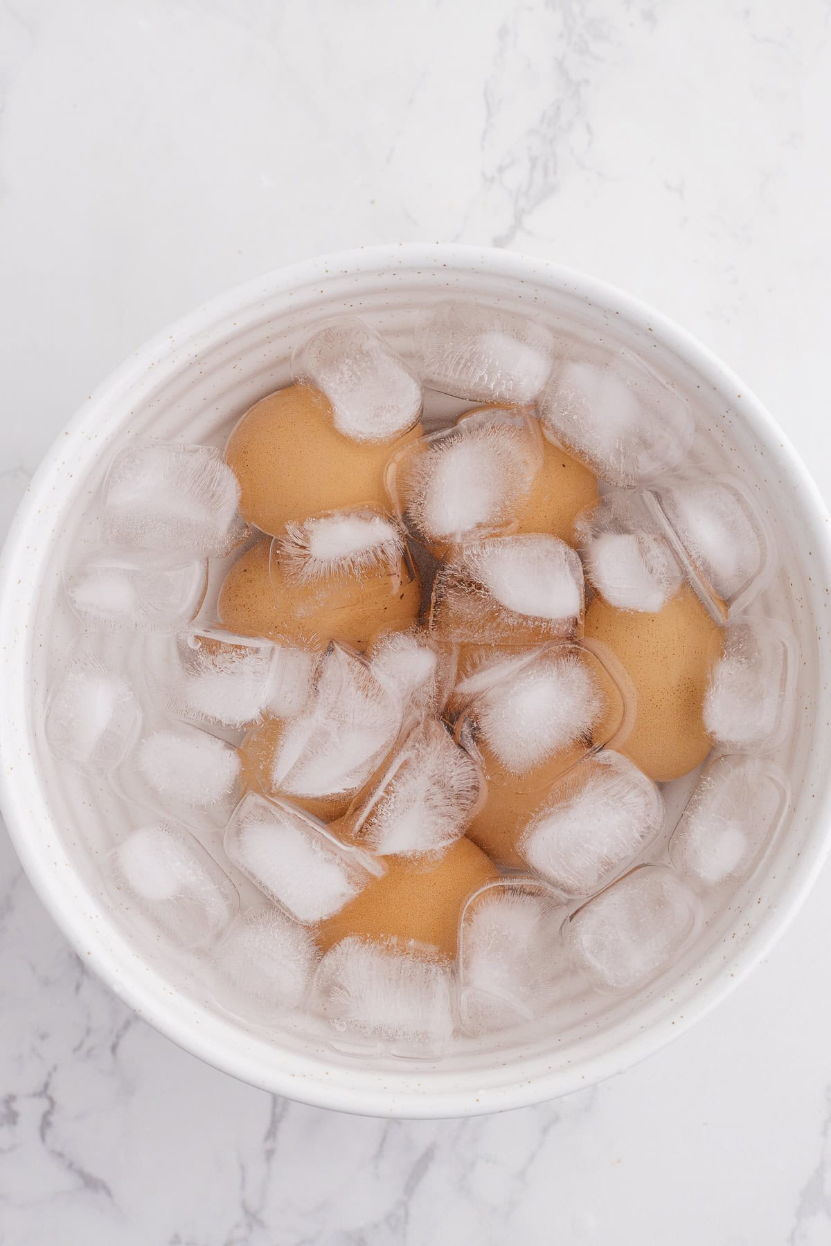 Brown eggs are submerged in a white bowl filled with ice water and ice cubes on a white marble surface.
