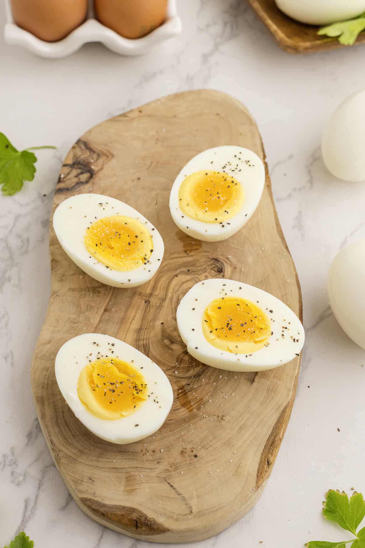 Four halved boiled eggs with slightly soft yolks are arranged on a wooden board, sprinkled with black pepper.