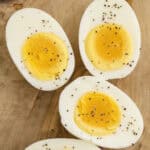 Four halved hard-boiled eggs are arranged on a wooden surface, with yolks exposed and sprinkled with salt and black pepper.