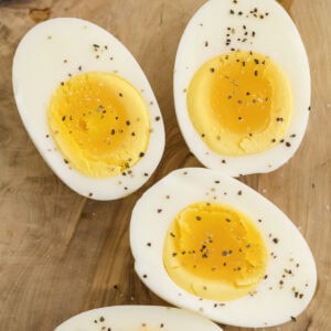 Four halved hard-boiled eggs are arranged on a wooden surface, with yolks exposed and sprinkled with salt and black pepper.