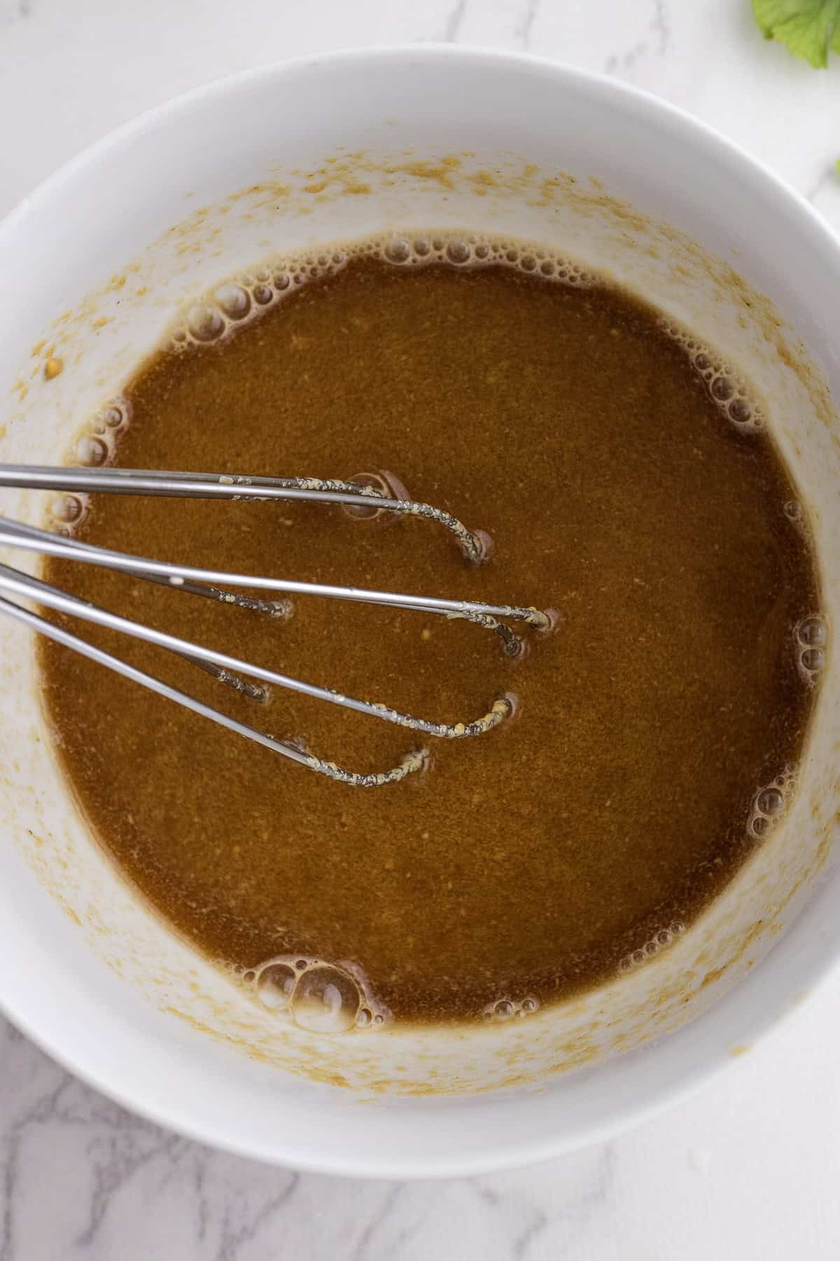A metal whisk in a white bowl mixing a brown liquid batter on a marble countertop.