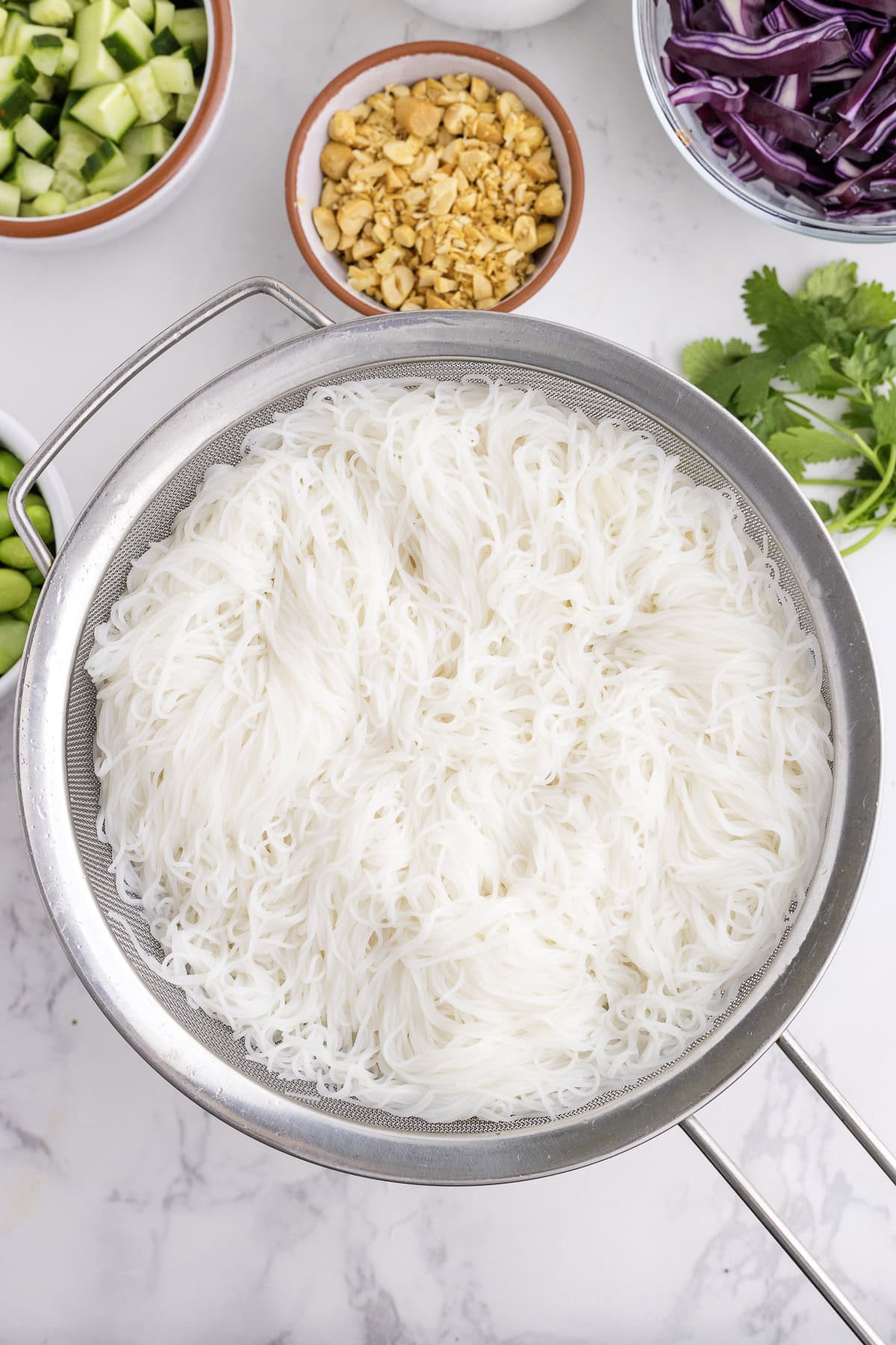 A metal strainer filled with cooked rice vermicelli noodles is surrounded by bowls of chopped peanuts, cucumber, purple cabbage, and fresh cilantro.