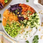 A bowl of rice noodles topped with edamame, shredded carrots, red cabbage, red bell peppers, cucumber, chopped peanuts, and fresh herbs, arranged in sections.