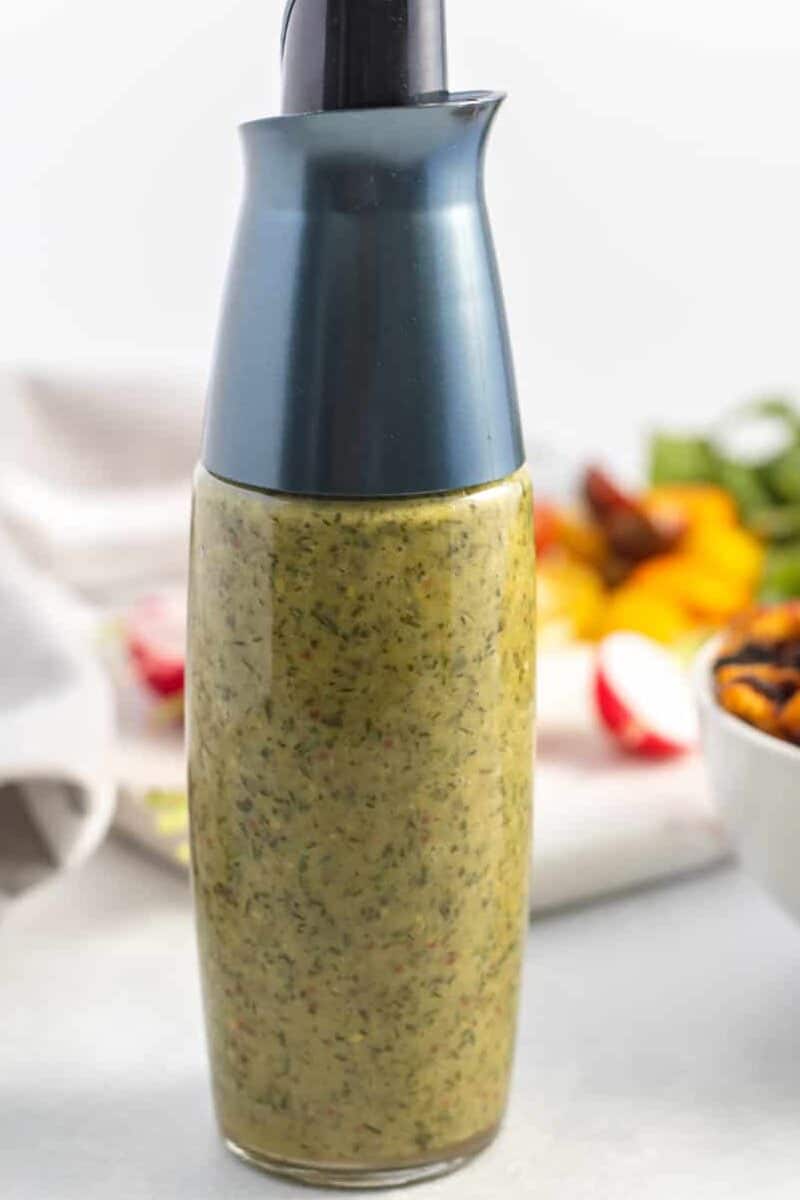 A clear glass bottle with a black cap filled with a green herb-speckled salad dressing, set on a light surface with blurred vegetables in the background.