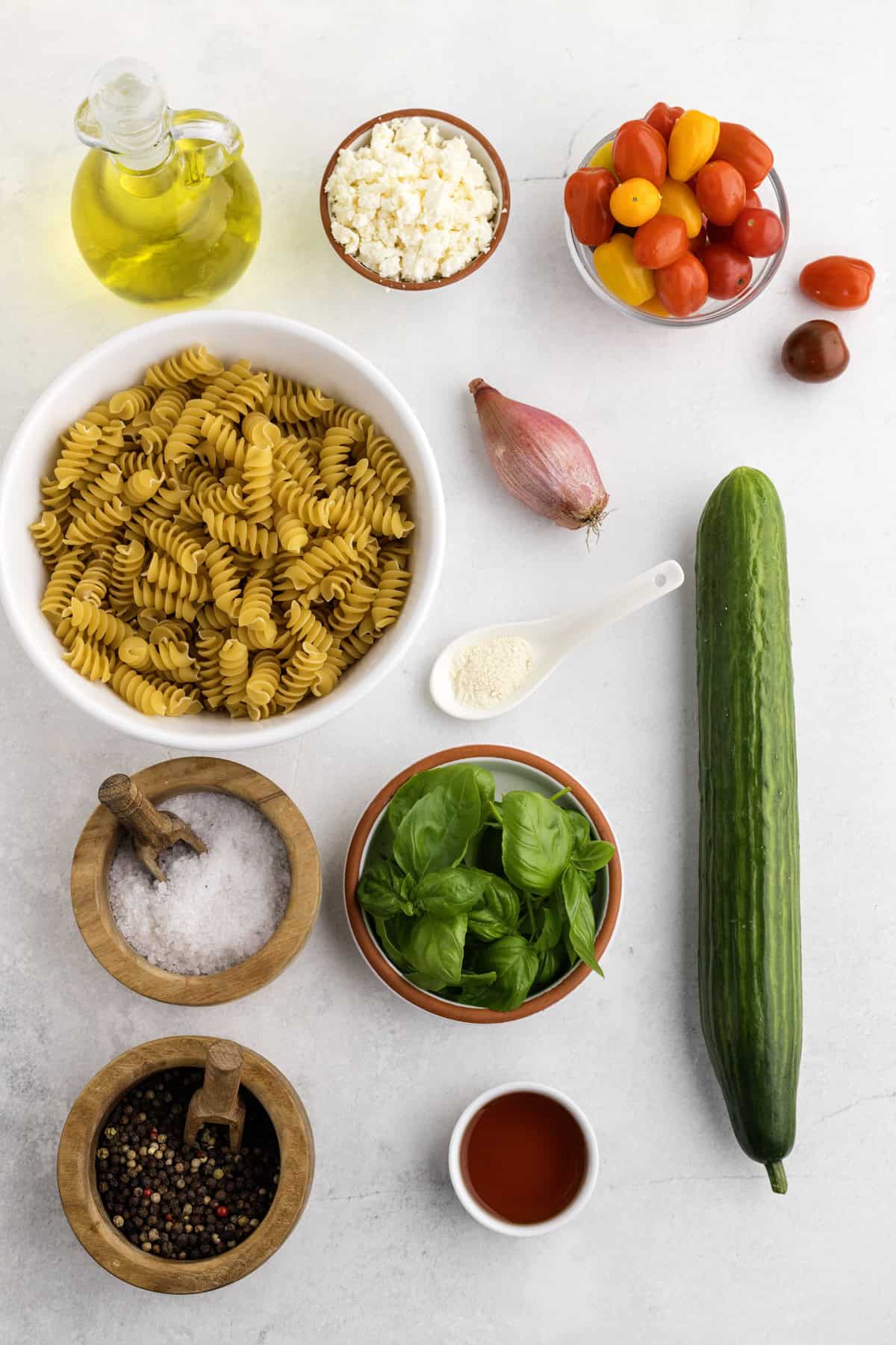 Ingredients for a pasta salad arranged on a white surface, including pasta, cucumber, tomatoes, feta cheese, shallot, basil, olive oil, seasonings, and vinegar.