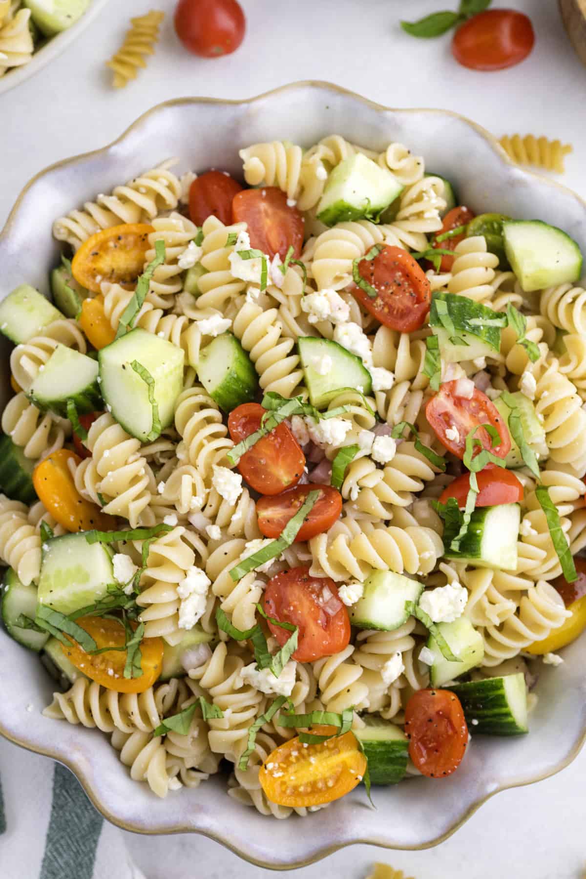 A bowl of rotini pasta salad with cherry tomatoes, cucumber, feta cheese, and fresh herbs.