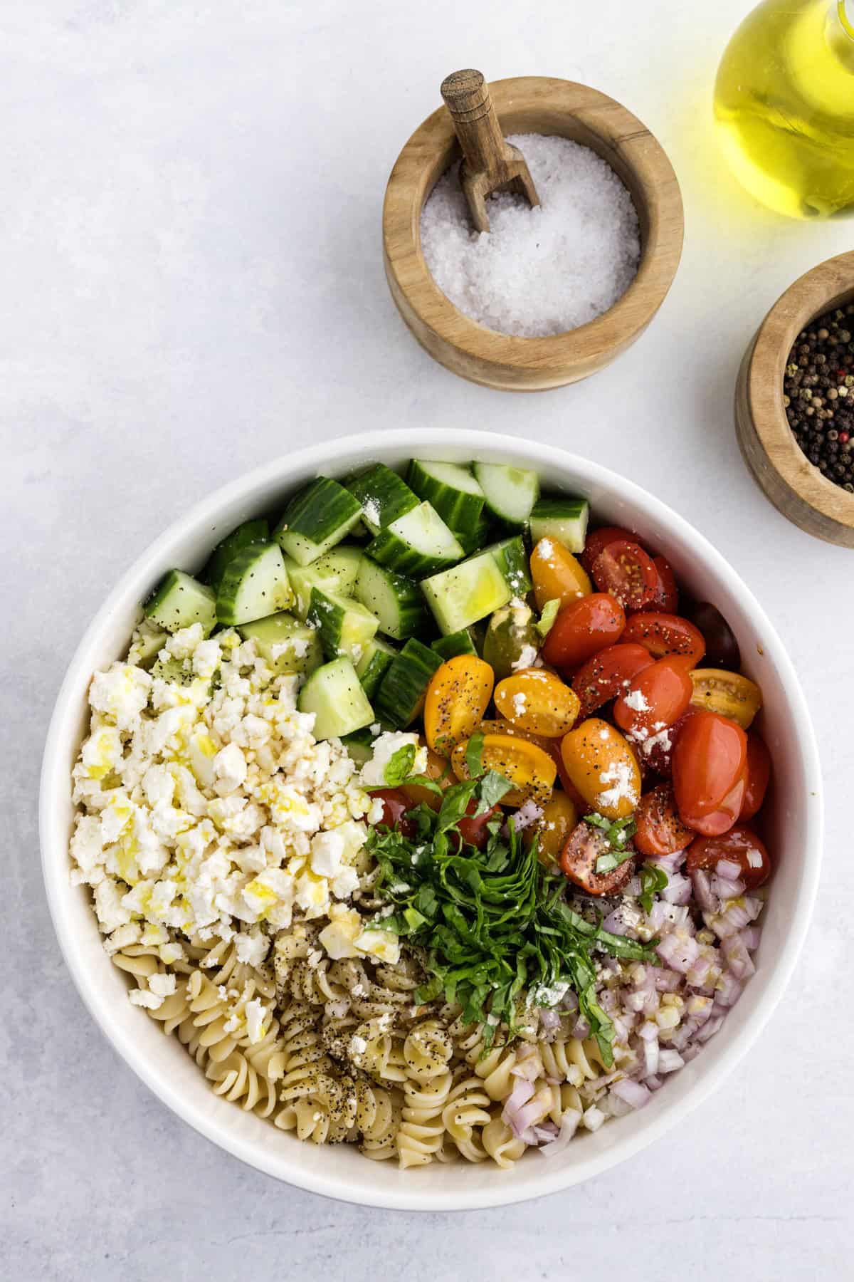 A bowl with cooked pasta, chopped cucumber, cherry tomatoes, feta cheese, red onion, and herbs, next to bowls of salt, pepper, and a bottle of olive oil on a light surface.