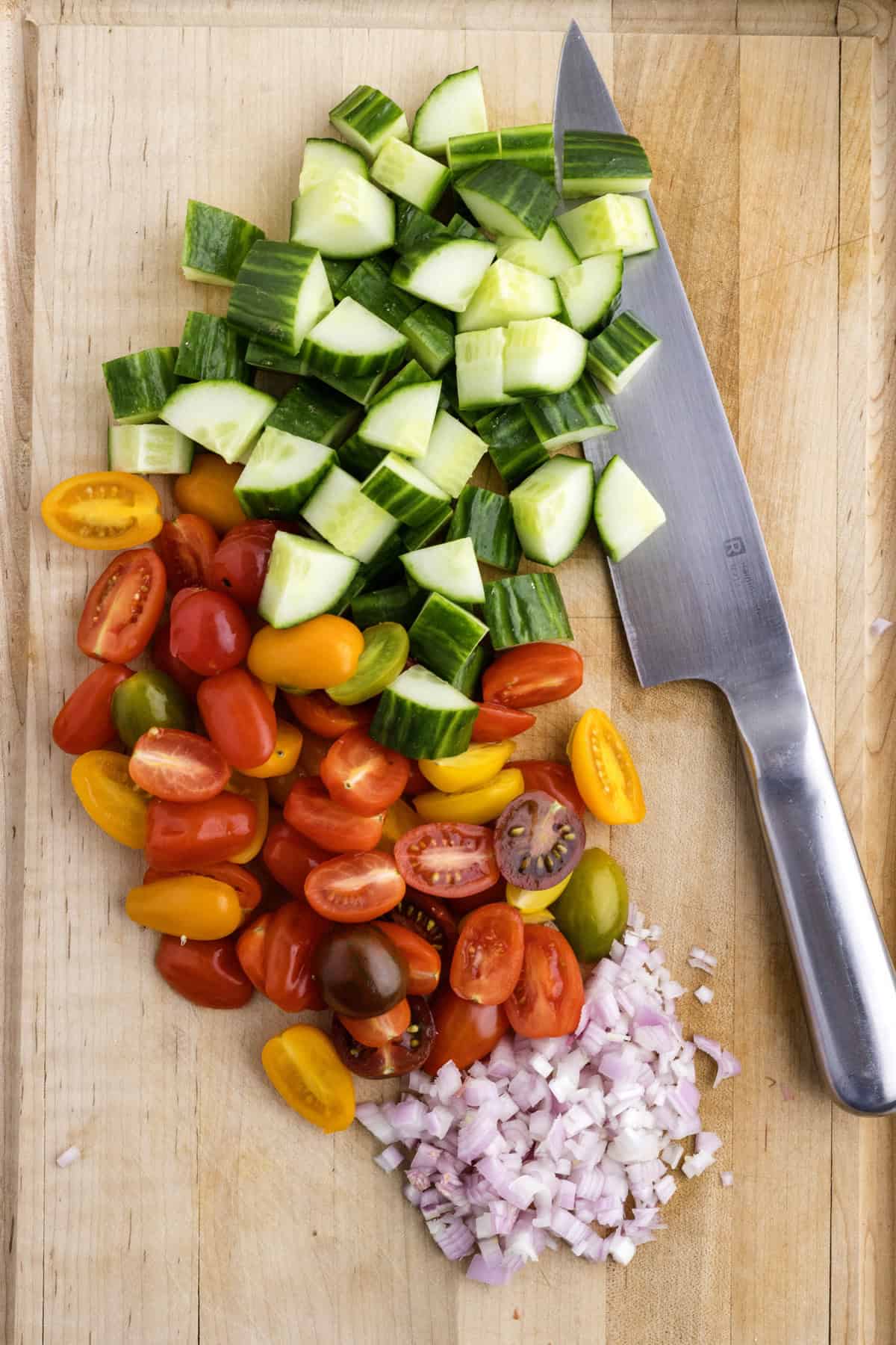 Chopped cucumber, cherry tomatoes, and shallots on a wooden cutting board with a knife.