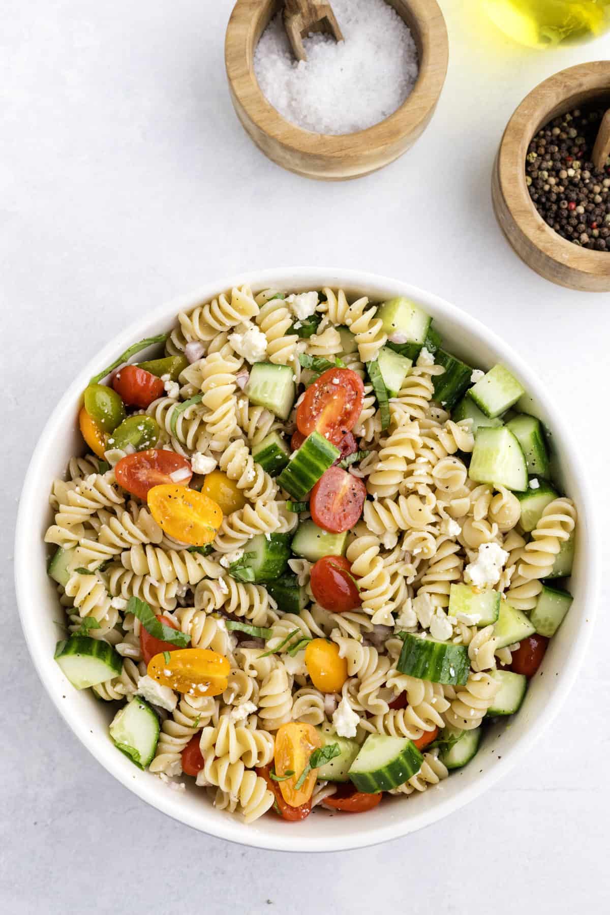 A bowl of rotini pasta salad with cherry tomatoes, cucumber, and feta cheese, next to wooden bowls of salt and pepper.
