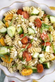 A bowl of cucumber and tomato pasta salad with cherry tomatoes, cucumber, feta cheese, and fresh herbs on a white surface.