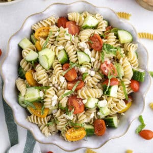 A bowl of cucumber and tomato pasta salad with cherry tomatoes, cucumber, feta cheese, and fresh herbs on a white surface.