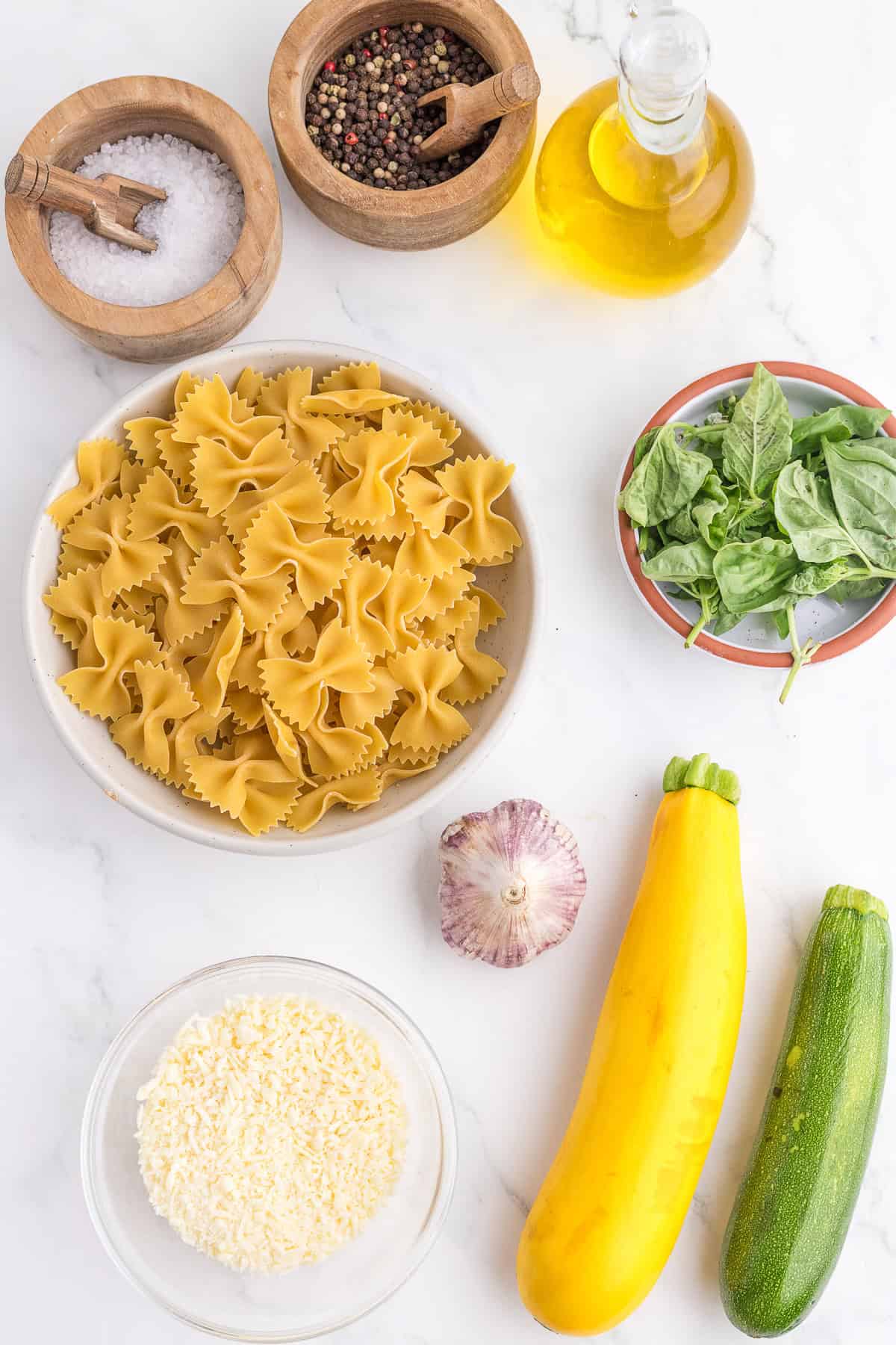 Bow-tie pasta, basil leaves, zucchini, yellow squash, garlic, grated cheese, salt, pepper, and olive oil arranged on a white surface.