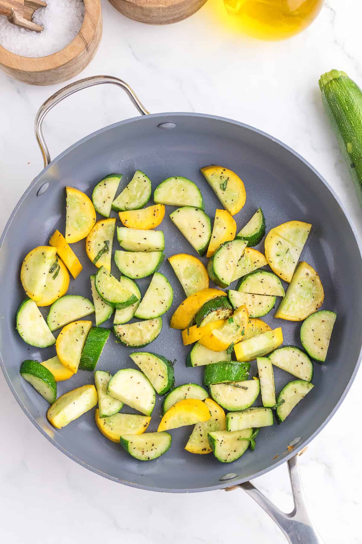 Sliced zucchini and yellow squash are sautéing in a gray frying pan on a marble countertop. Salt, pepper, and olive oil are visible nearby.
