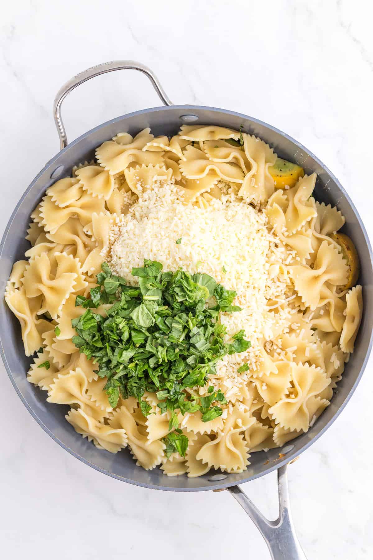 A skillet filled with cooked farfalle pasta, topped with shredded cheese and chopped fresh herbs on a marble surface.