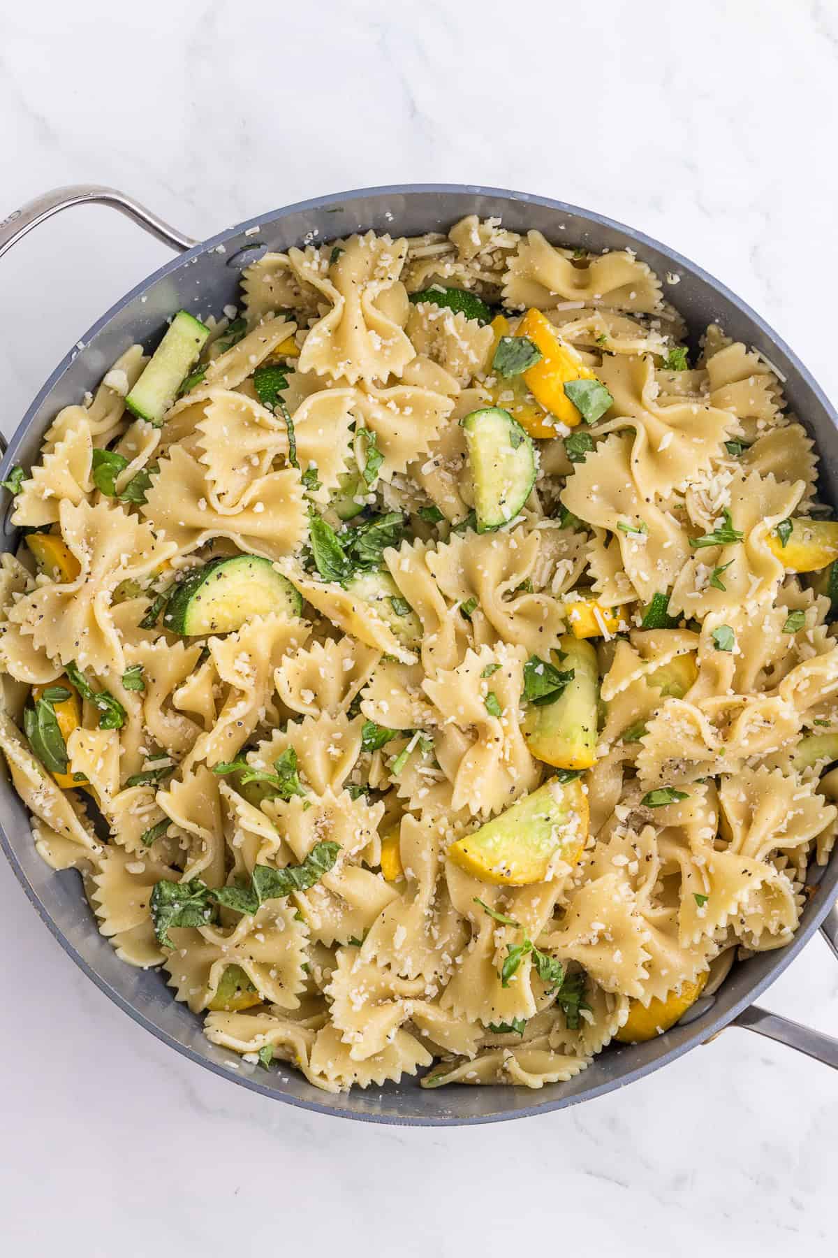 A pan filled with bowtie pasta mixed with sliced zucchini, yellow squash, herbs, and grated cheese on a white marble surface.