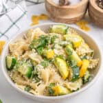 A bowl of farfalle pasta mixed with chopped zucchini, yellow squash, fresh herbs, and grated cheese sits on a white surface near wooden spice containers.