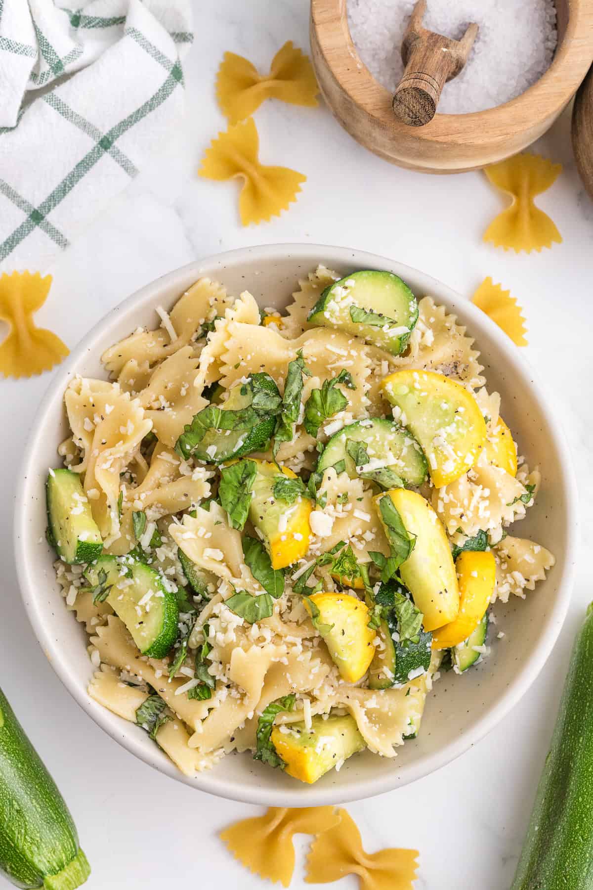 A bowl of farfalle pasta mixed with zucchini, yellow squash, fresh basil, and grated cheese, placed on a white surface with uncooked pasta and a salt container nearby.