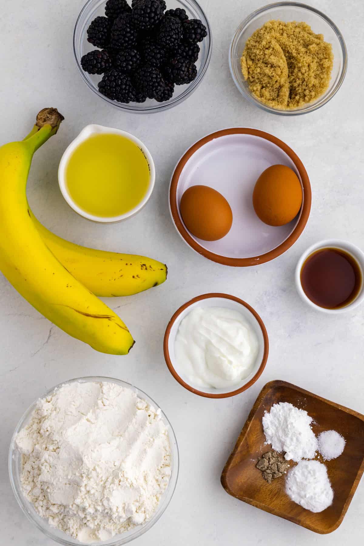 Top-down view of baking ingredients on a white surface, including bananas, blackberries, eggs, flour, brown sugar, yogurt, oil, vanilla extract, baking powder, and baking soda.