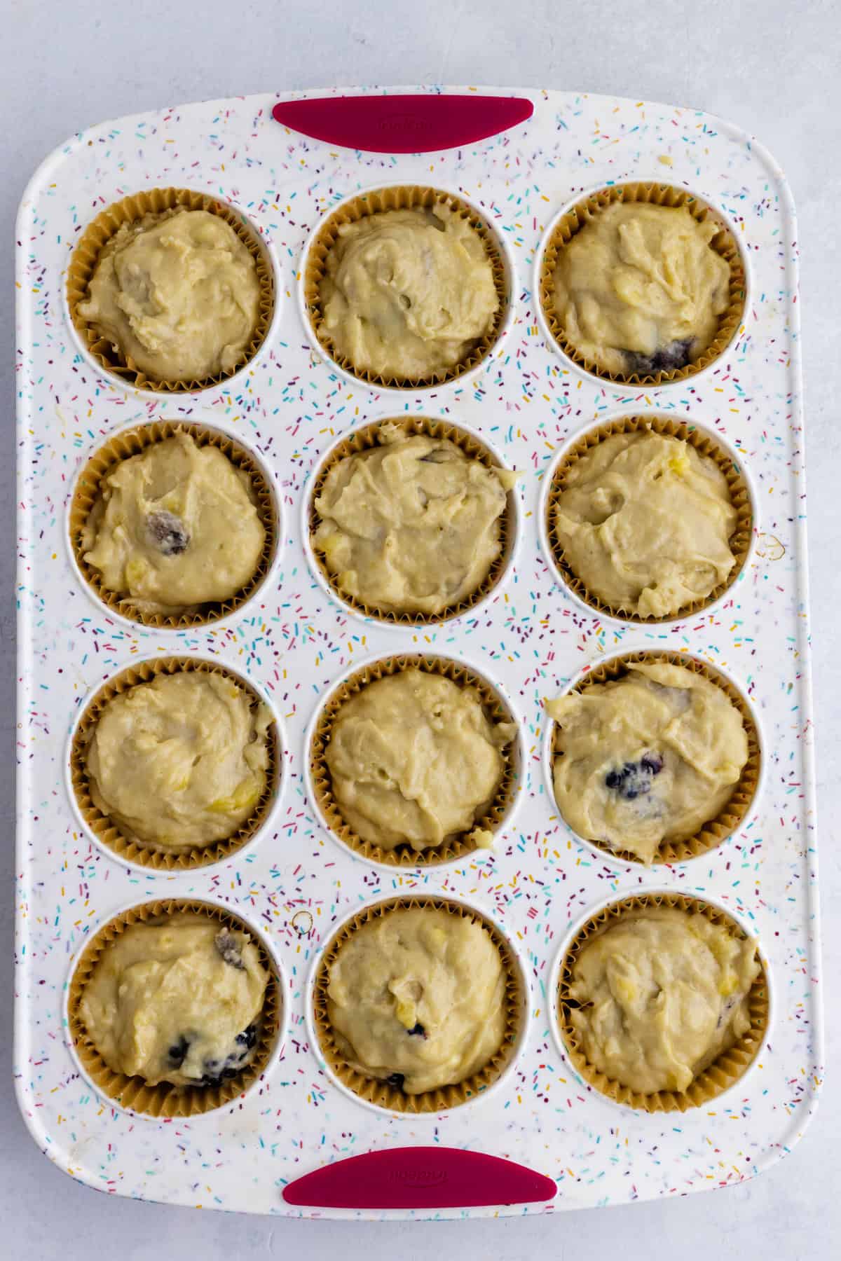 A muffin pan with 12 paper liners, each filled with unbaked muffin batter, sits on a light surface.
