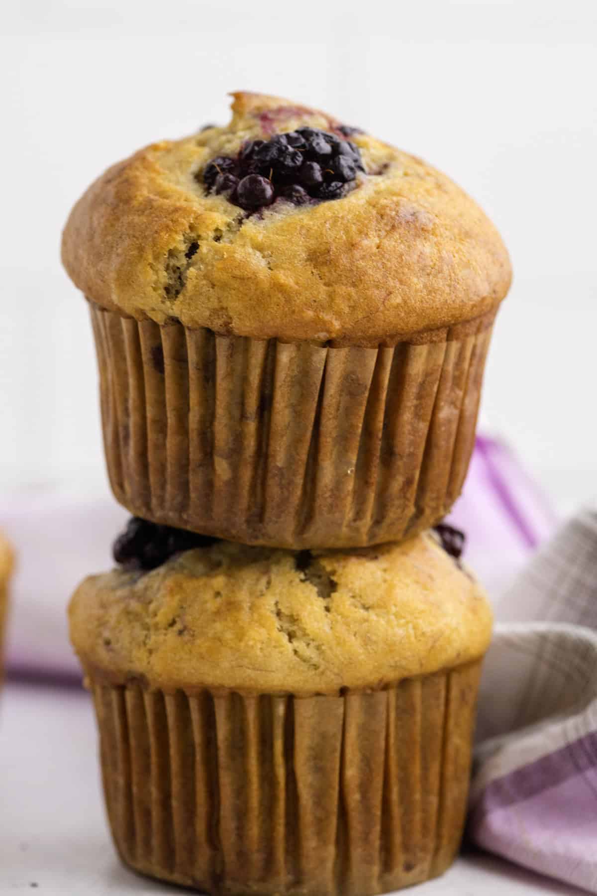 Two banana blackberry muffins stacked on top of each other, with visible blueberries on top, next to a folded plaid cloth.