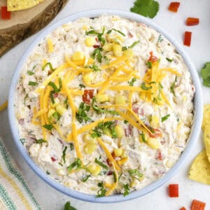 A bowl of creamy corn and cream cheese dip topped with shredded cheddar cheese, corn, chopped red peppers, and herbs; tortilla chips and cilantro garnish nearby.