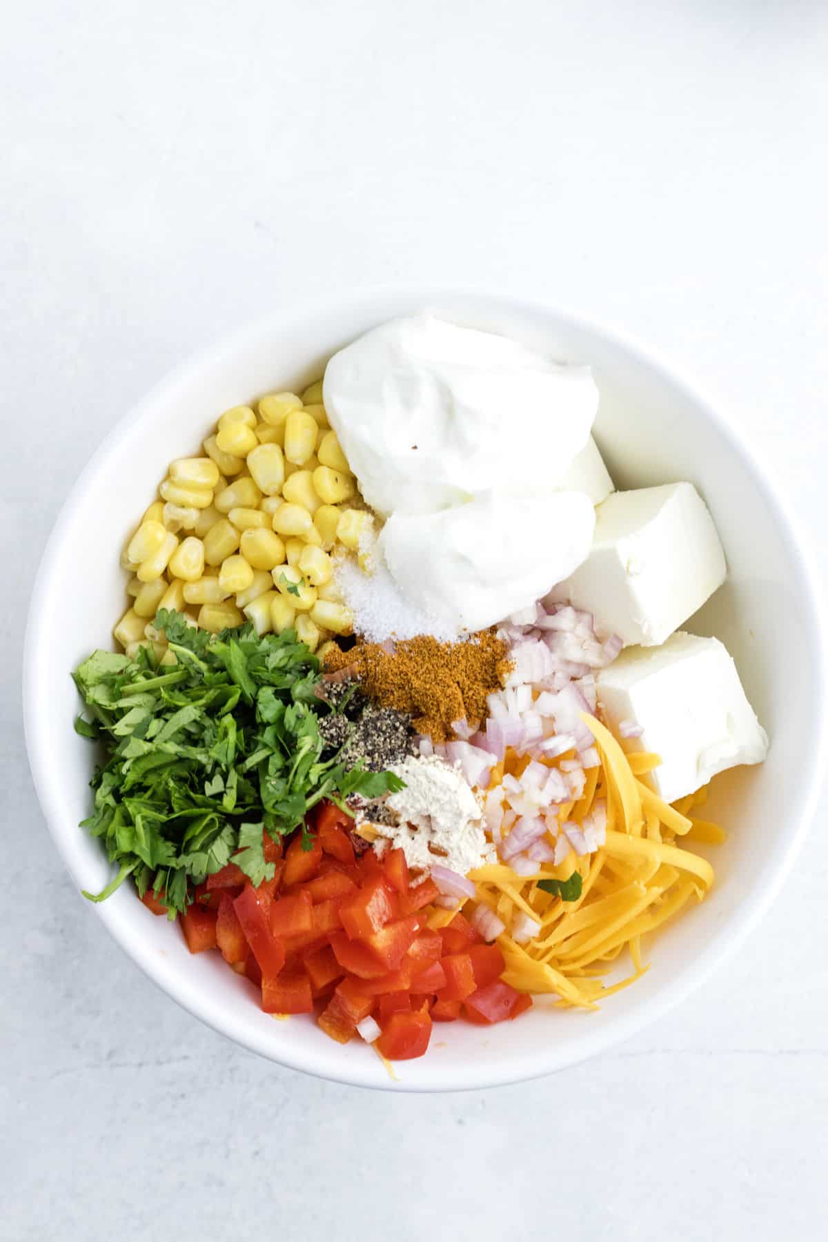 A white bowl containing corn, yogurt, cream cheese, shredded cheddar, chopped red bell pepper, red onion, cilantro, and spices, arranged in separate sections.