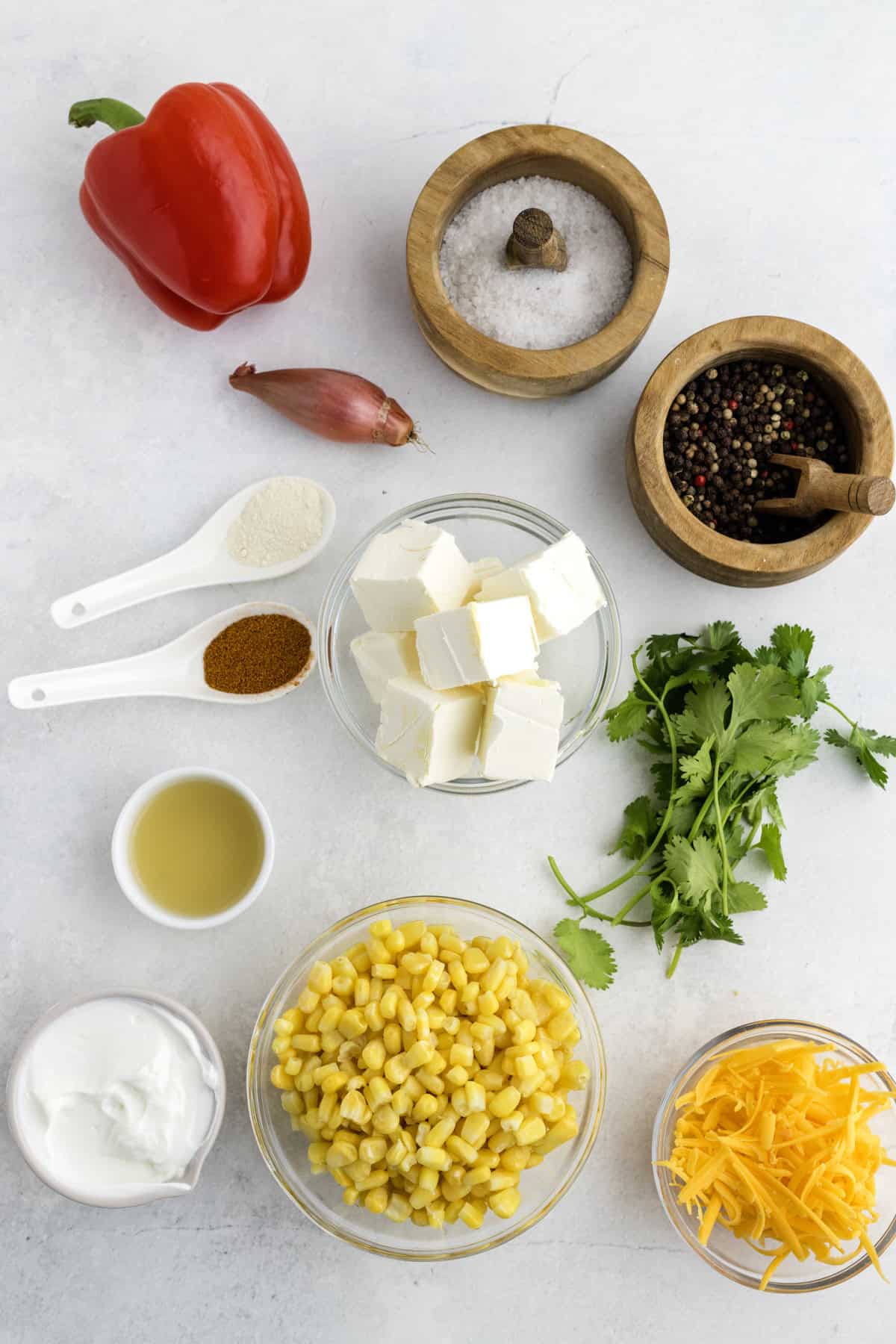 Assorted ingredients on a white surface including red bell pepper, corn, cream cheese, shredded cheese, yogurt, cilantro, spices, shallot, salt, pepper, and lime juice in small bowls.