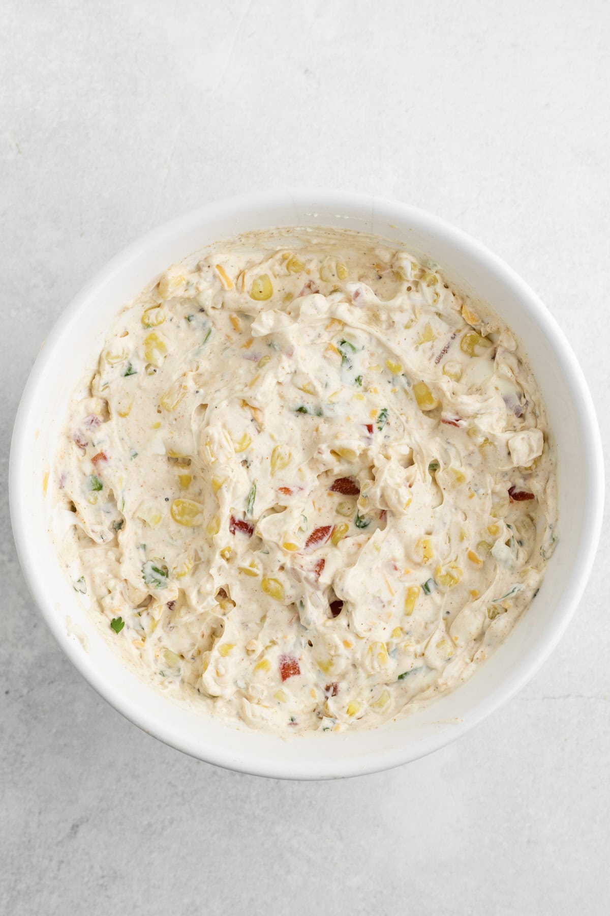 A white bowl filled with a creamy dip containing corn, red peppers, green herbs, and spices, placed on a light gray surface.