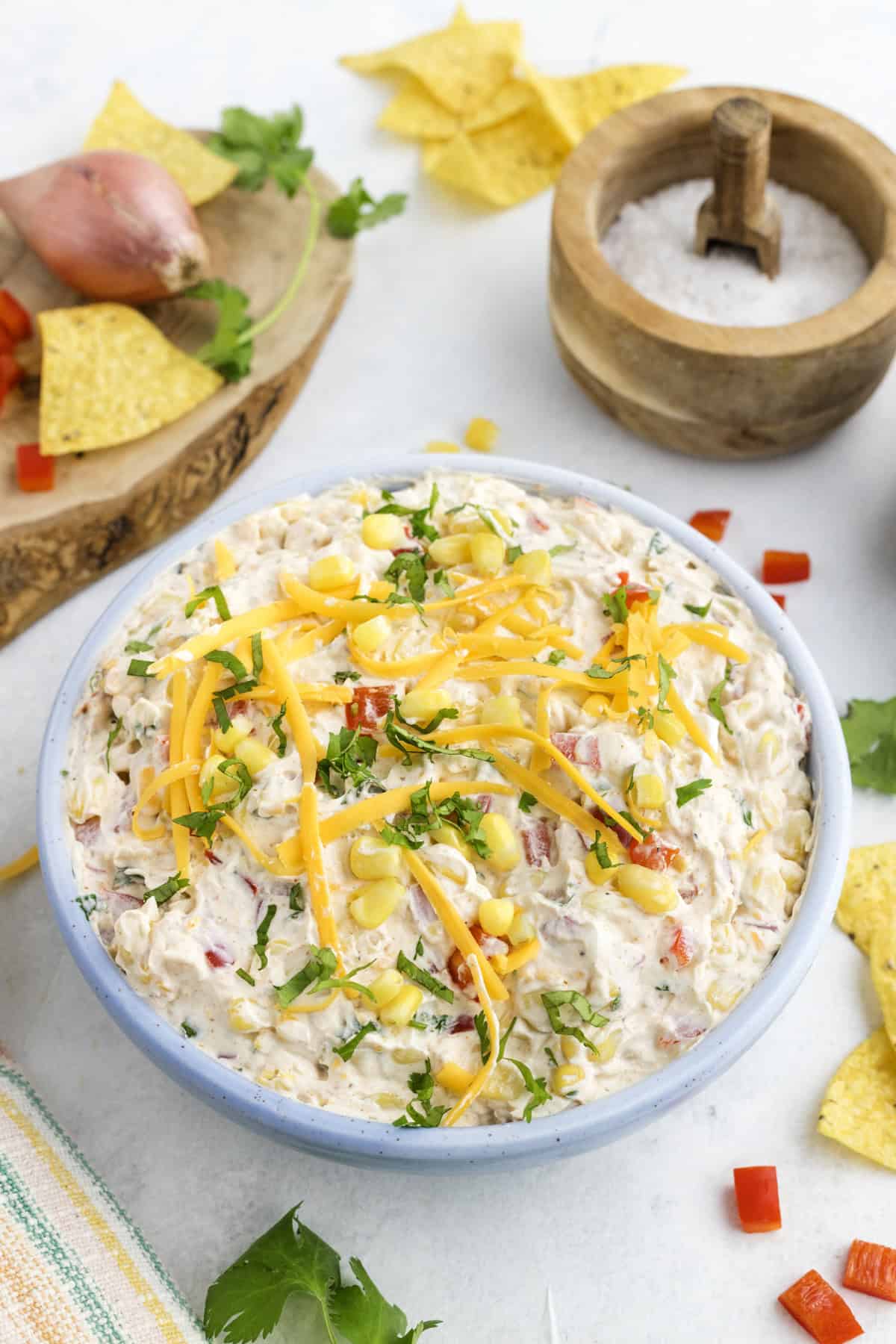 A bowl of creamy corn and cream cheese dip topped with shredded cheese, corn, and chopped herbs, surrounded by tortilla chips, cilantro, a salt bowl, and diced red pepper on a white surface.