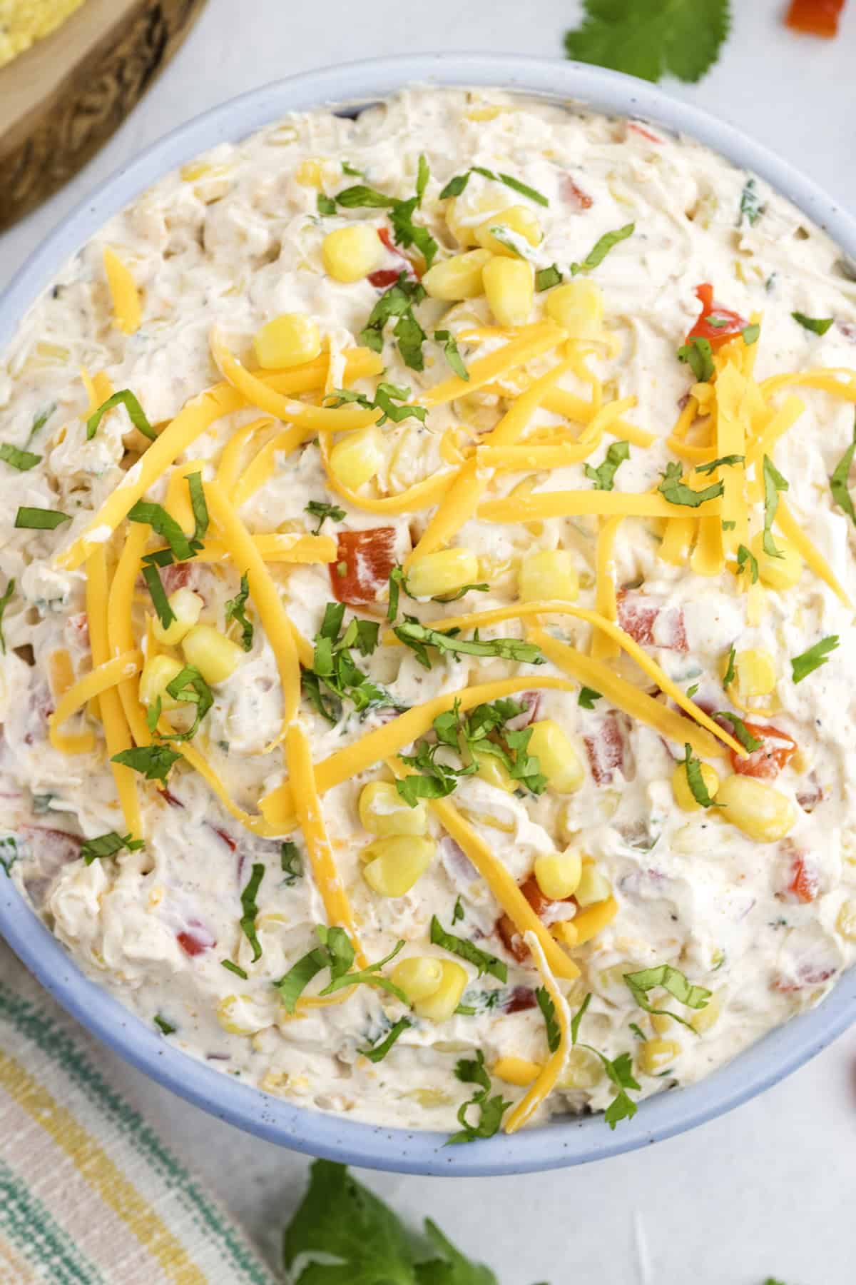 A bowl of creamy dip topped with shredded cheddar cheese, corn kernels, and chopped fresh herbs, with visible red pepper pieces mixed in.