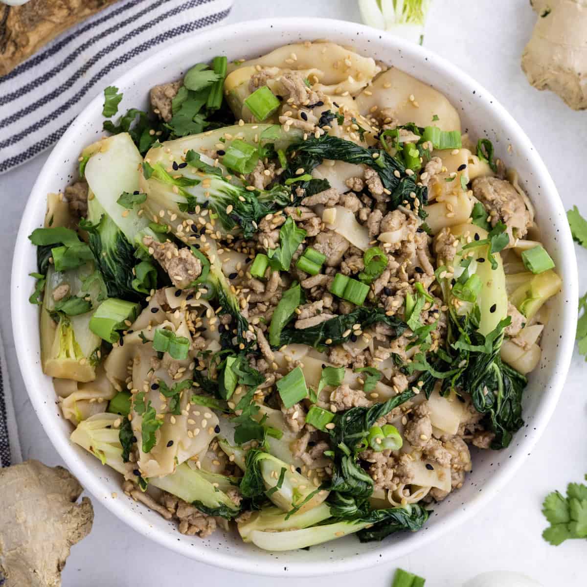 A white bowl filled with a Dumpling noodle dish featuring ground meat, bok choy, and garnishes like scallions, cilantro, and sesame seeds.