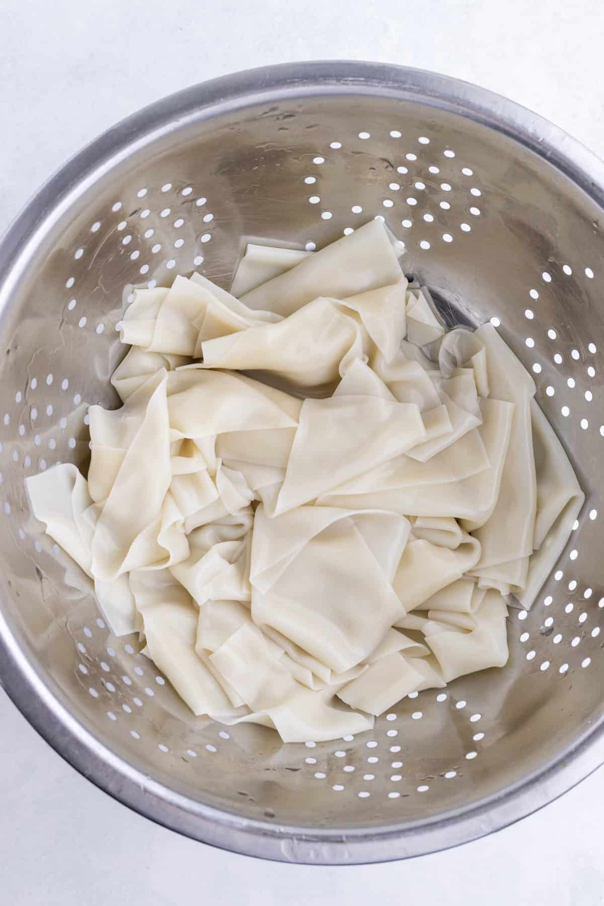 Wide, flat dumpling noodles are piled in a stainless steel colander.