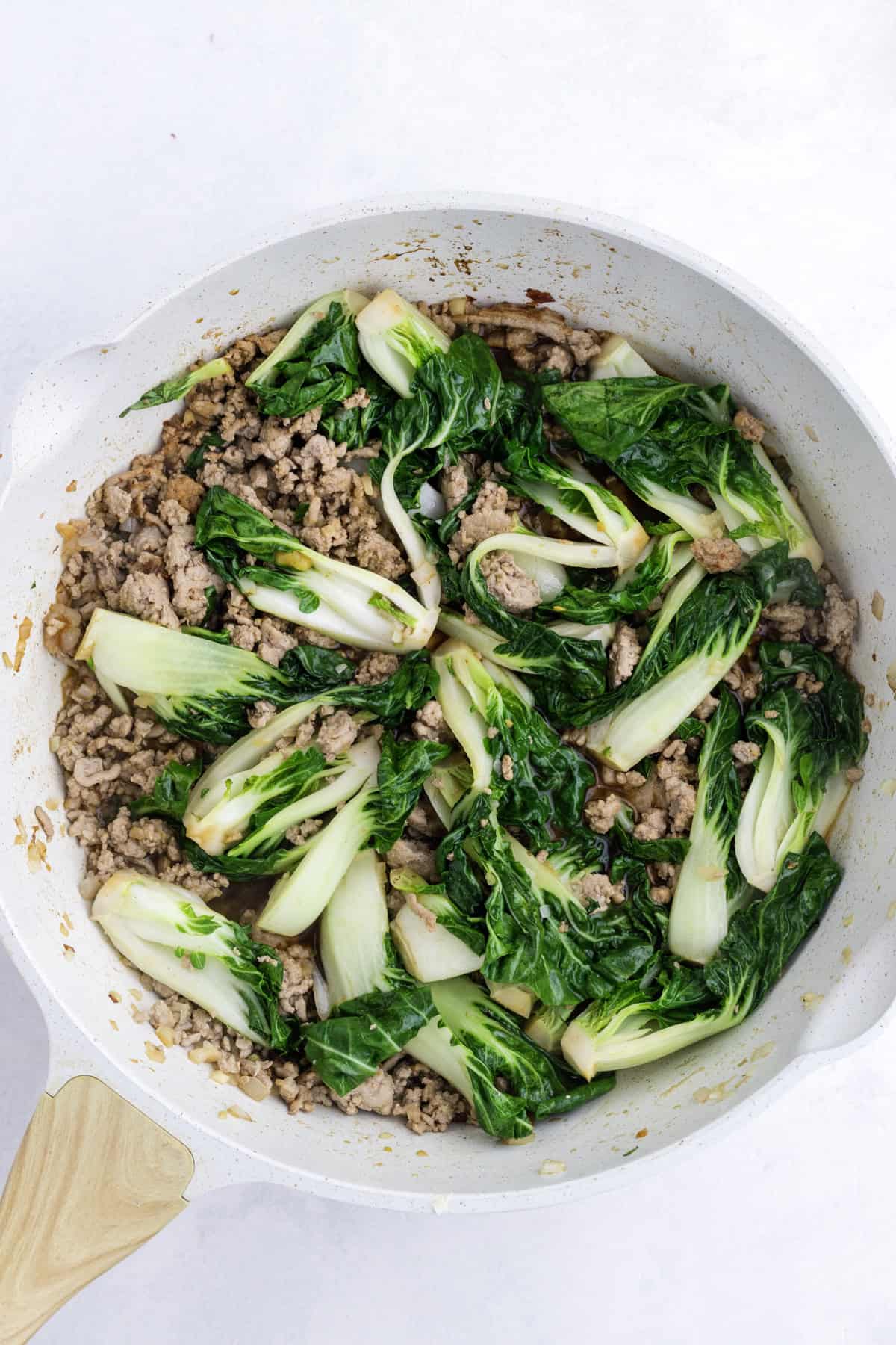 Ground meat and bok choy being cooked in a white pan.