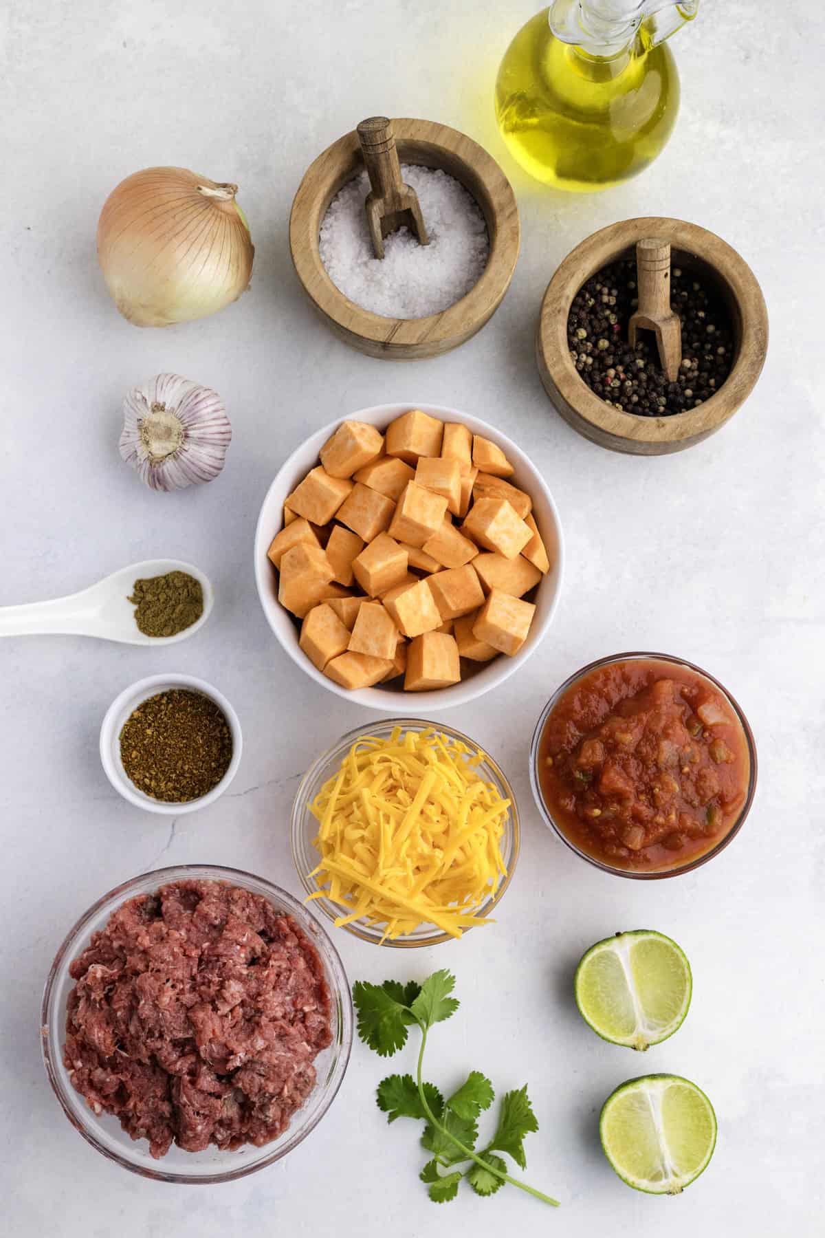 Ingredients laid out on a white surface, including ground beef, sweet potato cubes, shredded cheese, salsa, onion, garlic, spices, cilantro, lime halves, olive oil, salt, and pepper.