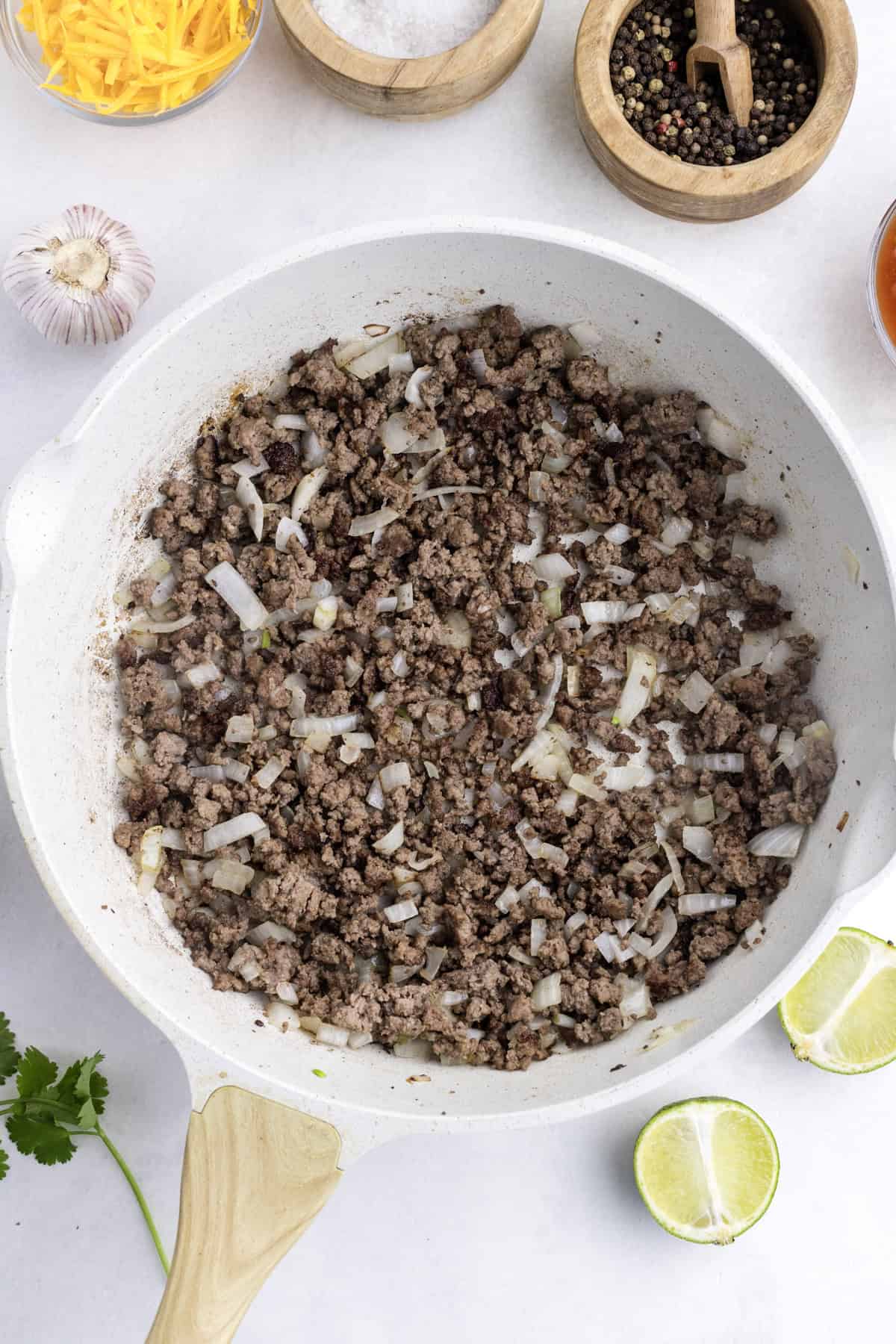 Ground beef and chopped onions cooking in a white skillet, surrounded by bowls of spices, garlic, shredded cheese, cilantro, and lime halves on a white surface.
