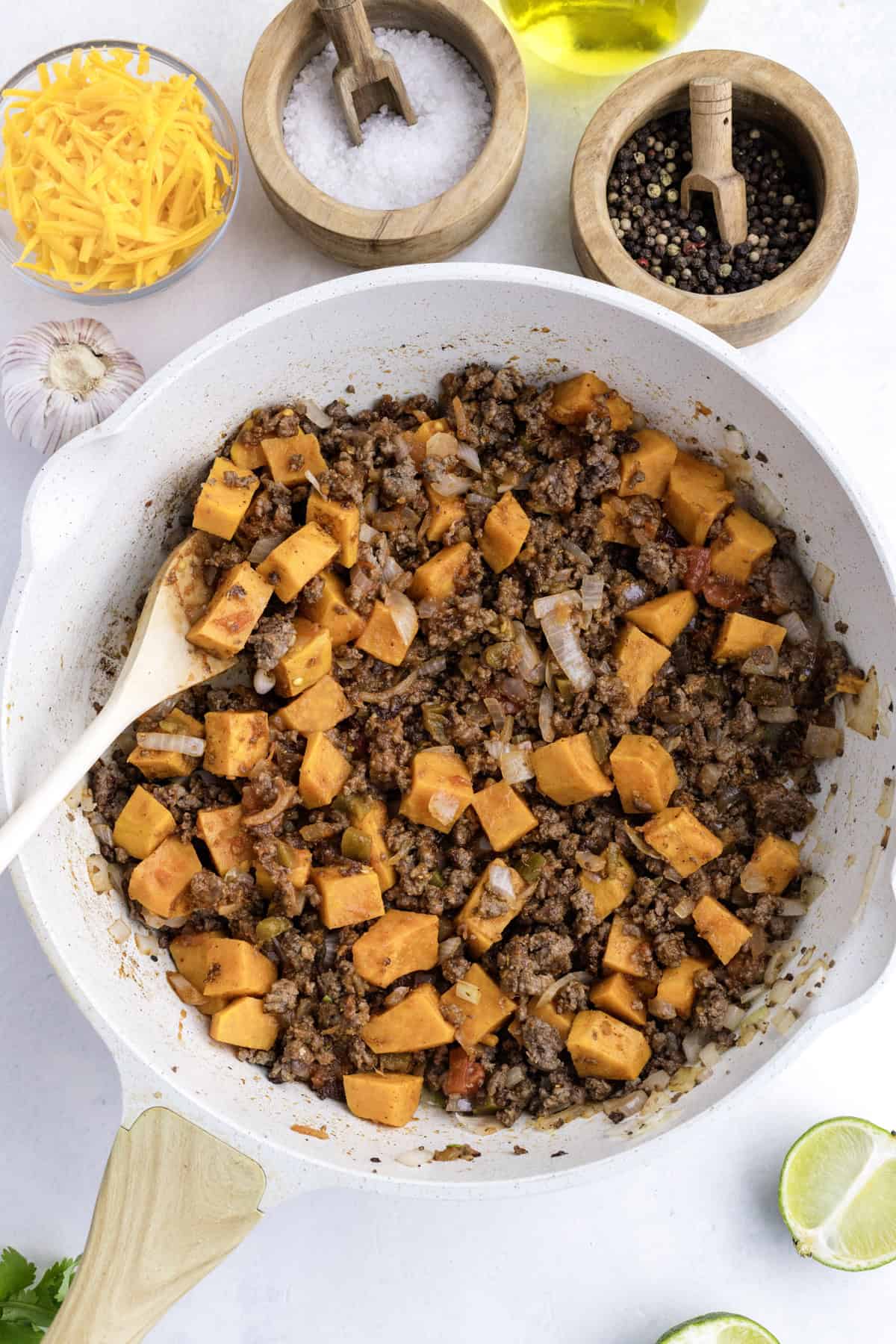 A skillet filled with cooked ground beef, diced sweet potatoes, and onions sits beside bowls of shredded cheese, salt, pepper, a bulb of garlic, olive oil, and a cut lime.