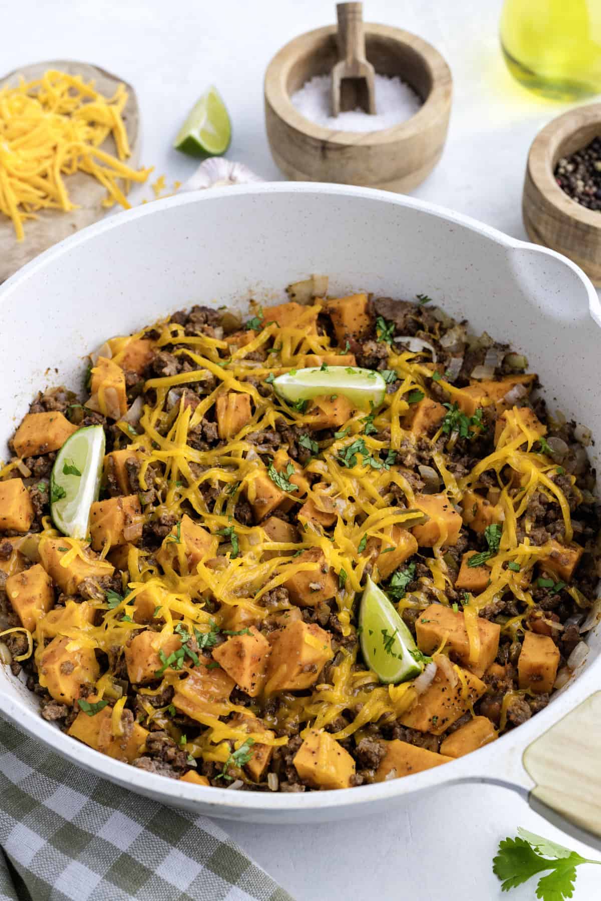 A skillet filled with ground beef, diced sweet potatoes, melted shredded cheese, herbs, and lime wedges, with salt, pepper, and olive oil containers nearby.