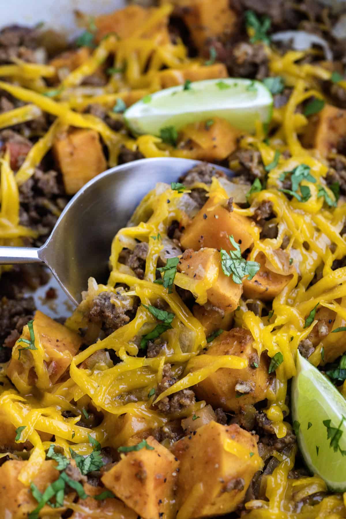 A close-up of a cheesy ground beef and sweet potato skillet topped with chopped cilantro and lime wedges, with a spoon serving a portion.