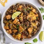 A bowl of ground beef hash with sweet potatoes, melted cheese, diced vegetables, chopped cilantro, and a lime wedge on top.