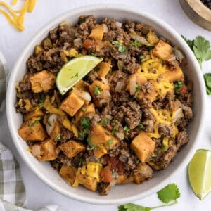 A bowl of ground beef hash with sweet potatoes, melted cheese, diced vegetables, chopped cilantro, and a lime wedge on top.