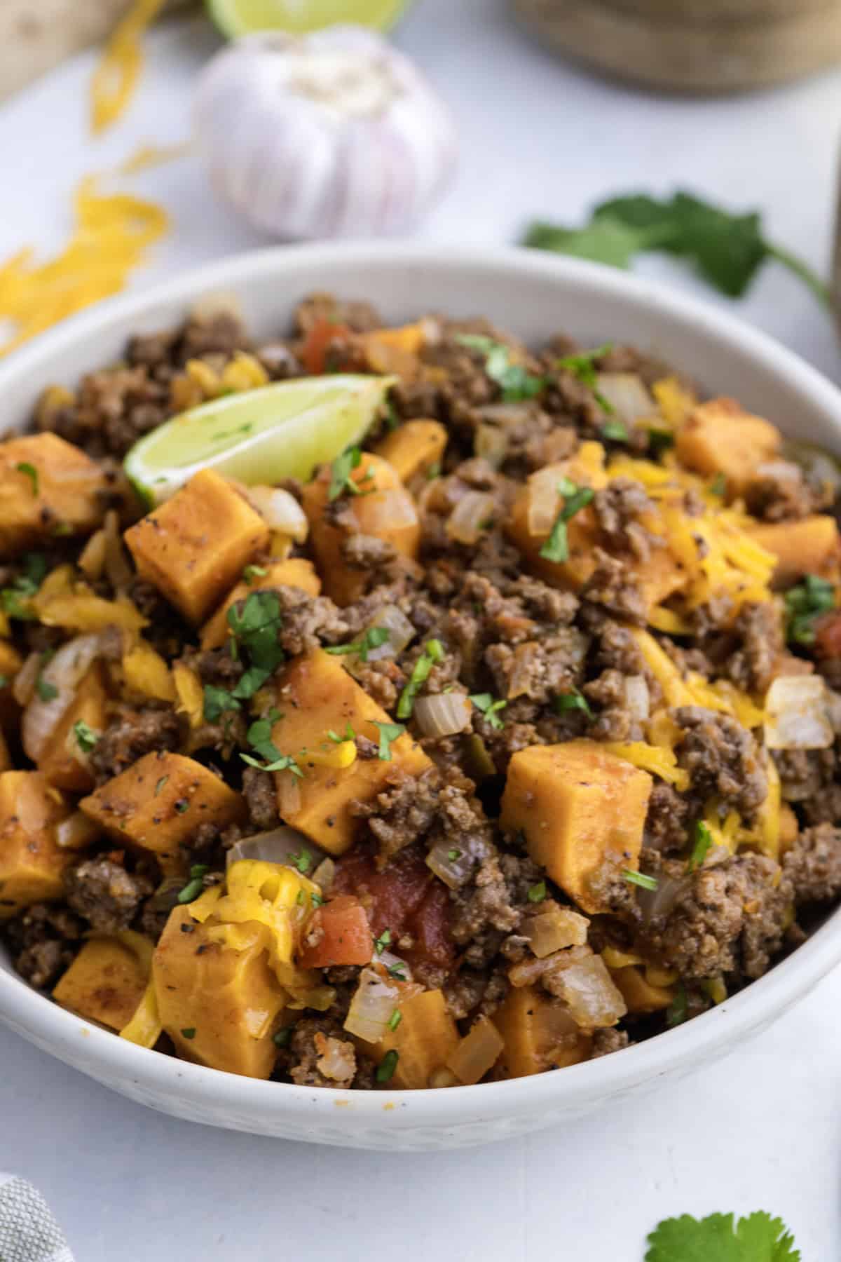 A bowl filled with a mixture of ground beef, diced sweet potatoes, onions, tomatoes, shredded cheese, and chopped herbs, garnished with a lime wedge.
