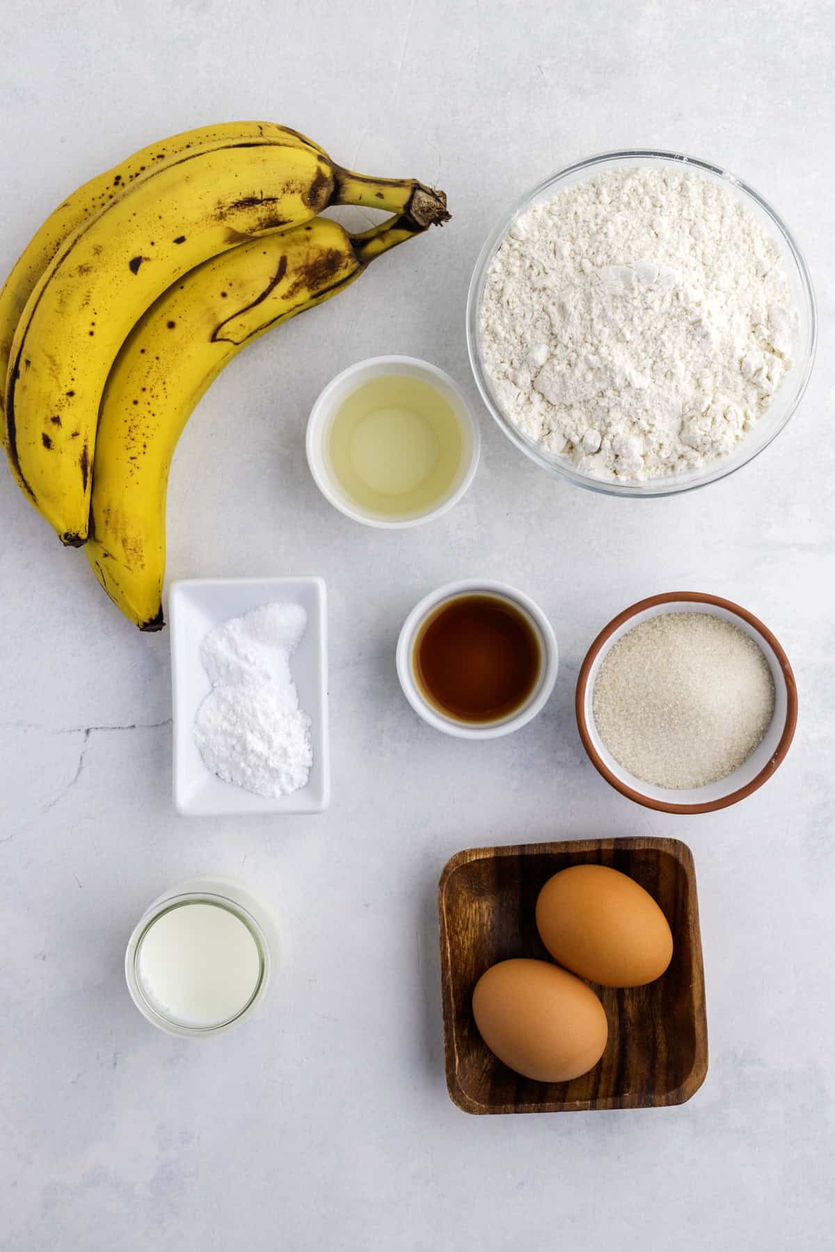 Bananas, flour, sugar, two eggs, baking powder, milk, vanilla extract, milk, vanilla and oil are arranged separately on a white surface.