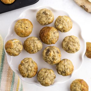 A white plate holds twelve small banana muffins arranged in a loose circle. More muffins and a striped cloth are partially visible nearby.