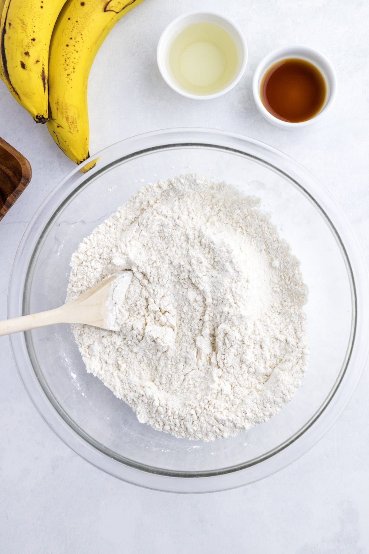 A glass bowl with flour mixture and a wooden spoon, next to bananas, a small bowl of oil, and a small bowl of vanilla extract on a white surface.