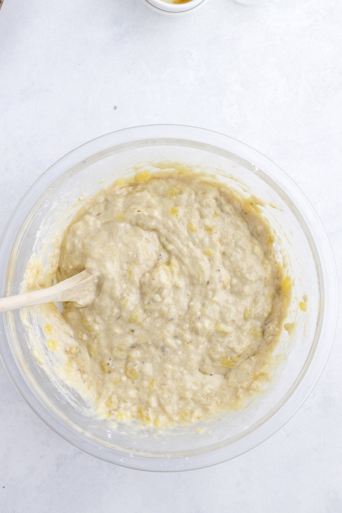 Clear glass bowl with banana bread batter being mixed by a wooden spoon on a light surface.