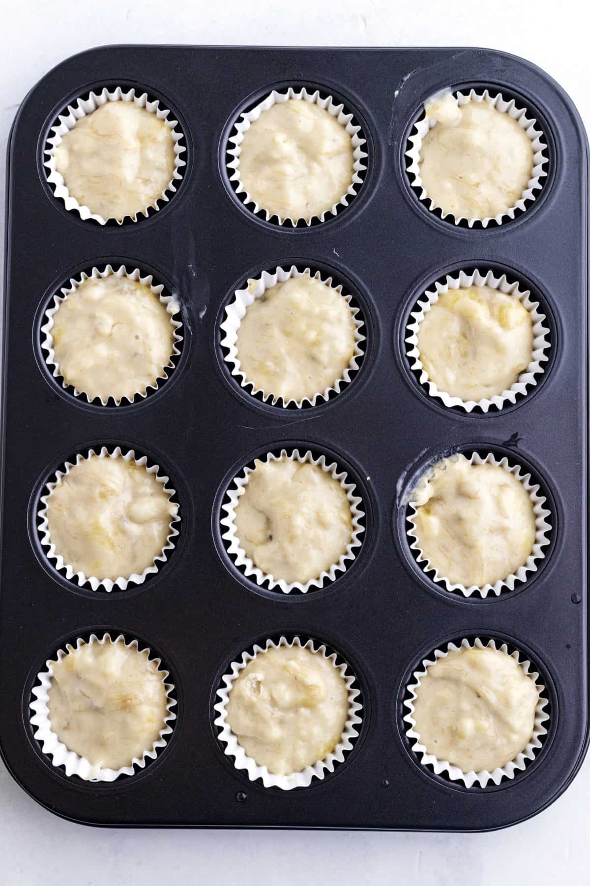 A muffin tin lined with white paper cups, each filled with unbaked batter, ready to be baked.