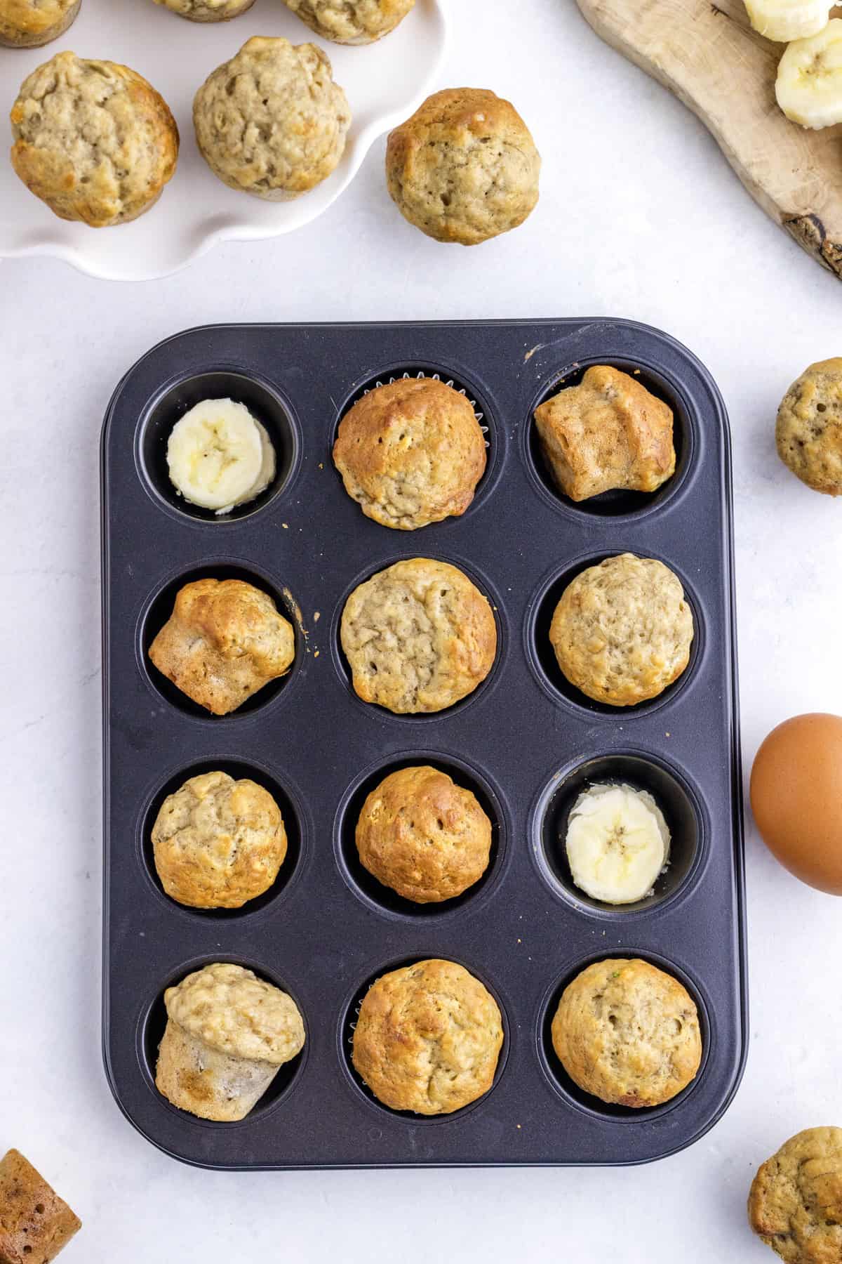 A muffin tin with eleven baked banana muffins and one compartment containing banana slices, surrounded by more muffins, an egg, and banana slices on a light surface.
