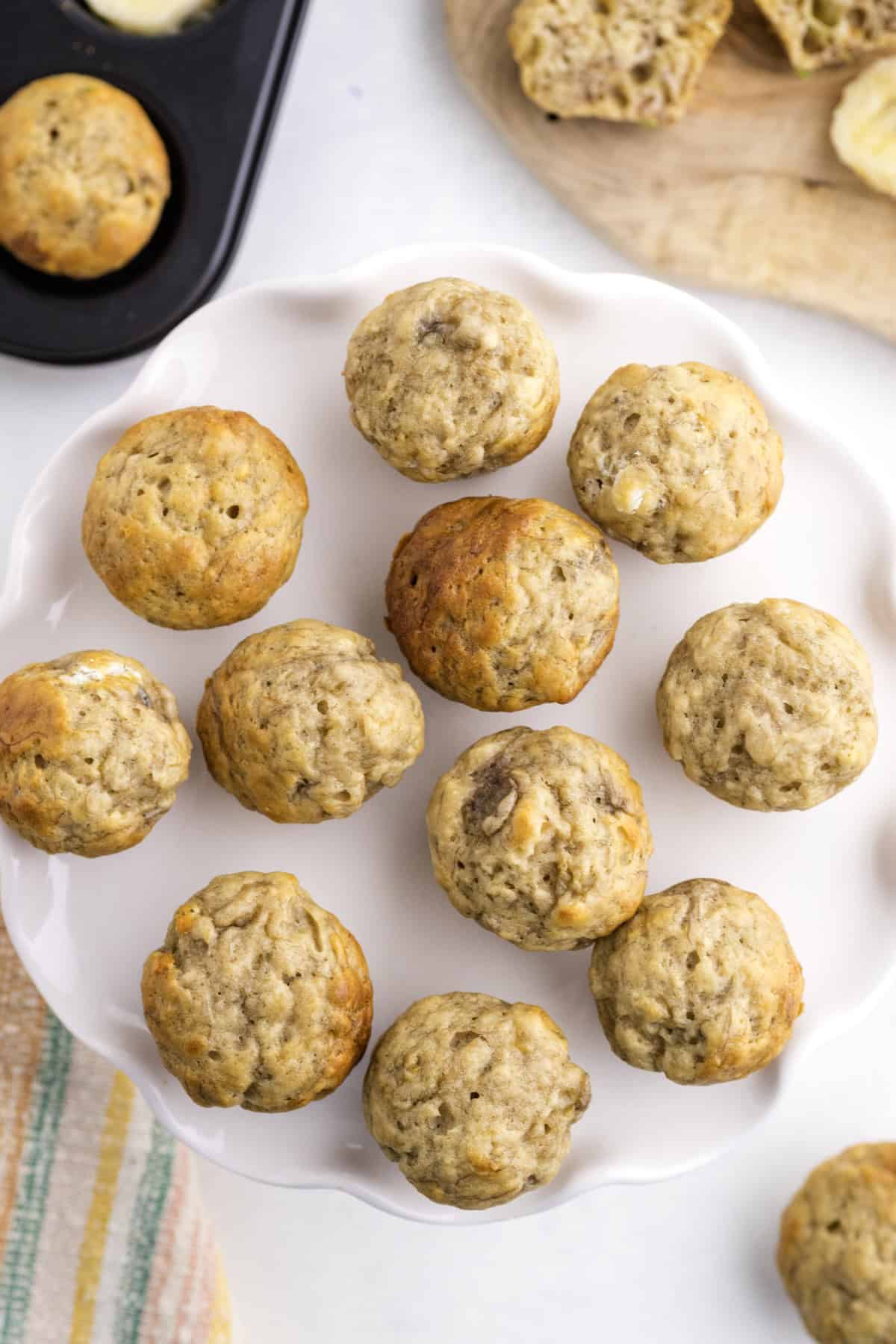 A white plate with twelve banana muffins, with a muffin tin and sliced bread visible in the background.
