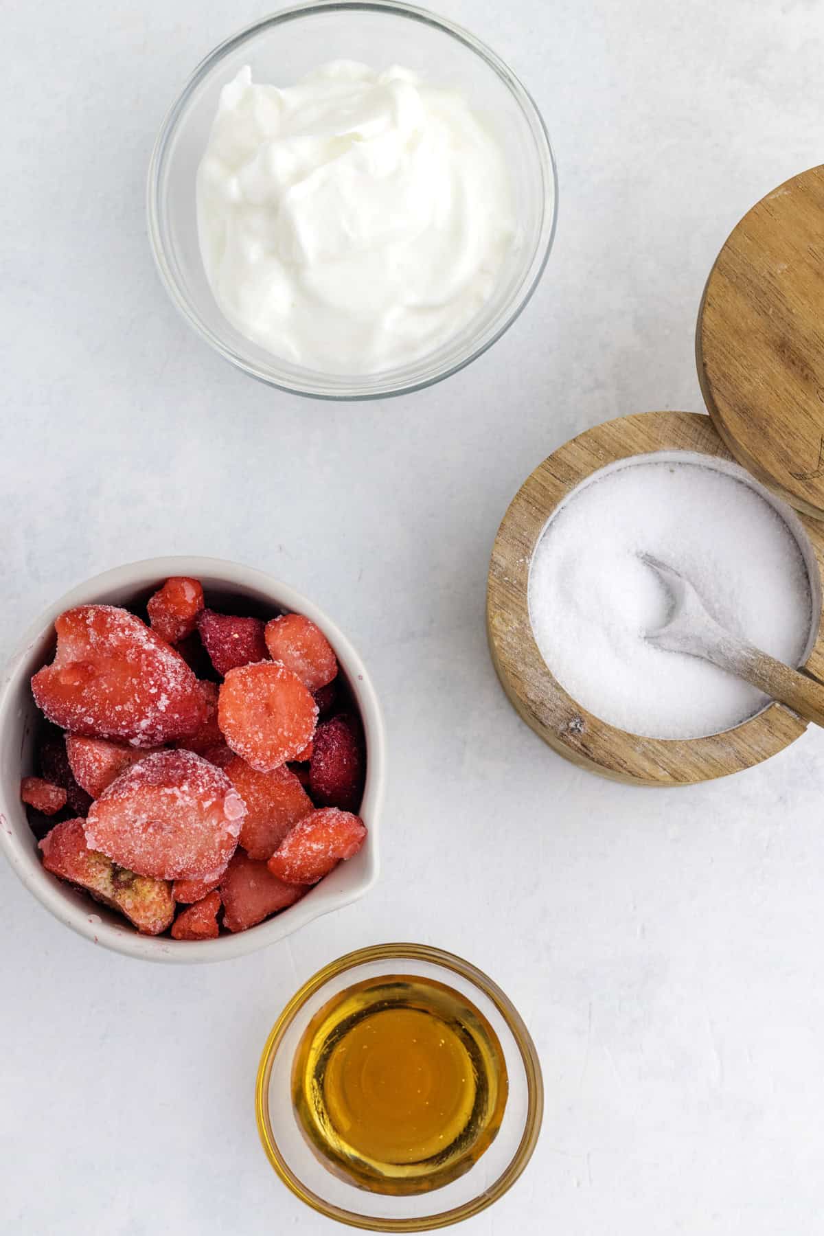 Un bol de fraises congelées, un bol de yaourt, une petite coupelle en verre contenant du sirop d'érable et un récipient en bois ouvert contenant du sucre sur une surface blanche.